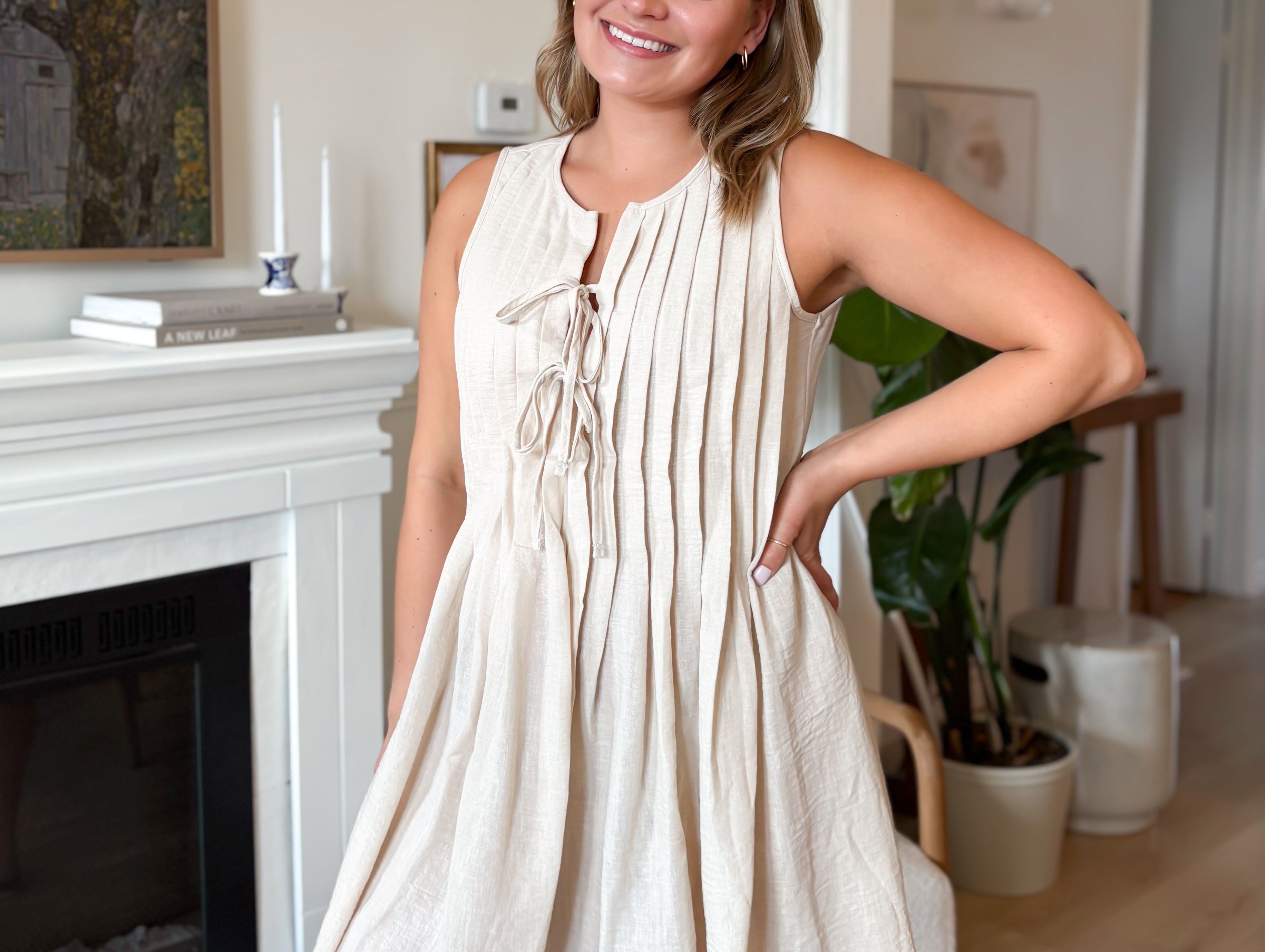 Woman in a beige dress standing in a living room with a fireplace and decor.